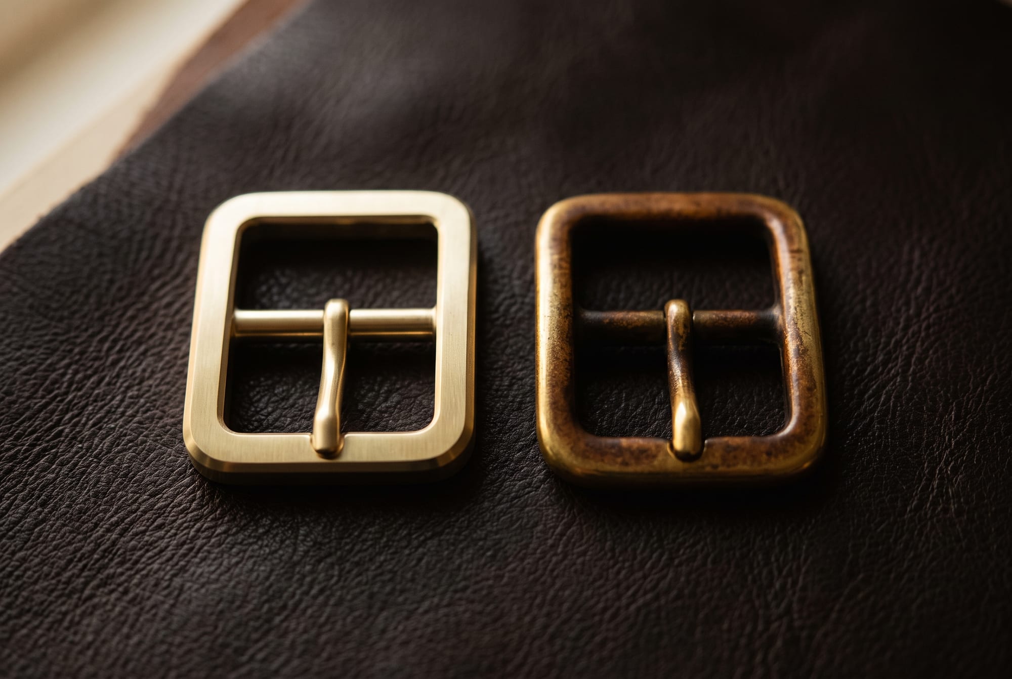 Two solid brass buckles side by side on dark leather, one bright and newly polished, the other deeply patinated to a warm mottled amber-brown from years of handling