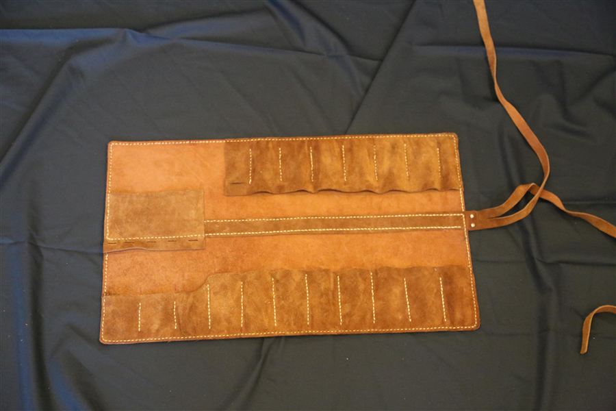 A rolled crazy horse leather tool and brush organiser, its pockets holding brushes, lying open on a work surface in warm light