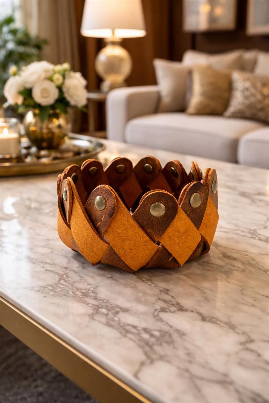 A woven cognac leather catchall bowl with brass rivets sitting on a white marble coffee table with a brass frame, a brass tray and white roses behind it