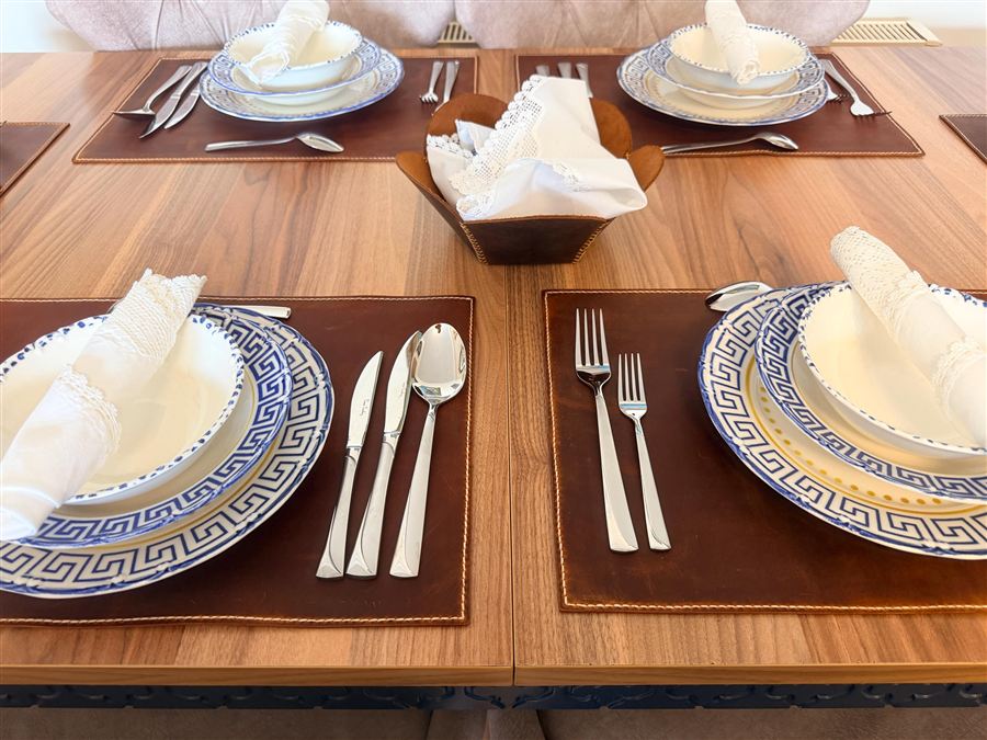 Four hand-stitched tobacco brown Crazy Horse leather placemats set on a walnut dining table with blue-and-white plates, cutlery and a matching leather bread basket